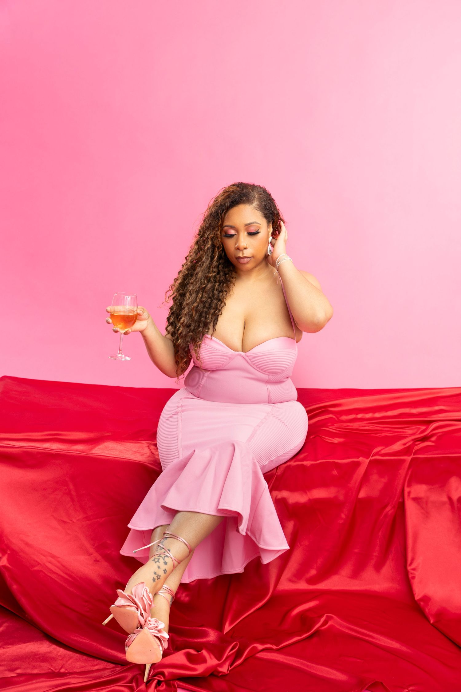 A curvy woman in a pink ruffled dress holds a wine glass while seated on red satin, against a pink background.