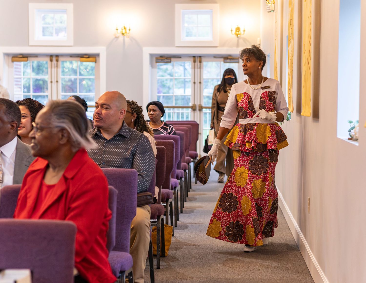 Congregation members in colorful attire gather in a church hall with purple pews during a religious service.