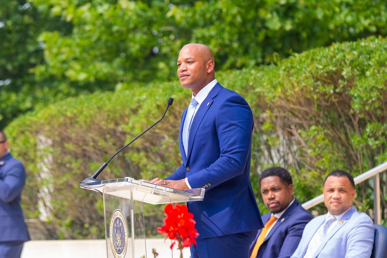 Man in blue suit speaking at podium during outdoor ceremony with red ribbon and seated guests in background.