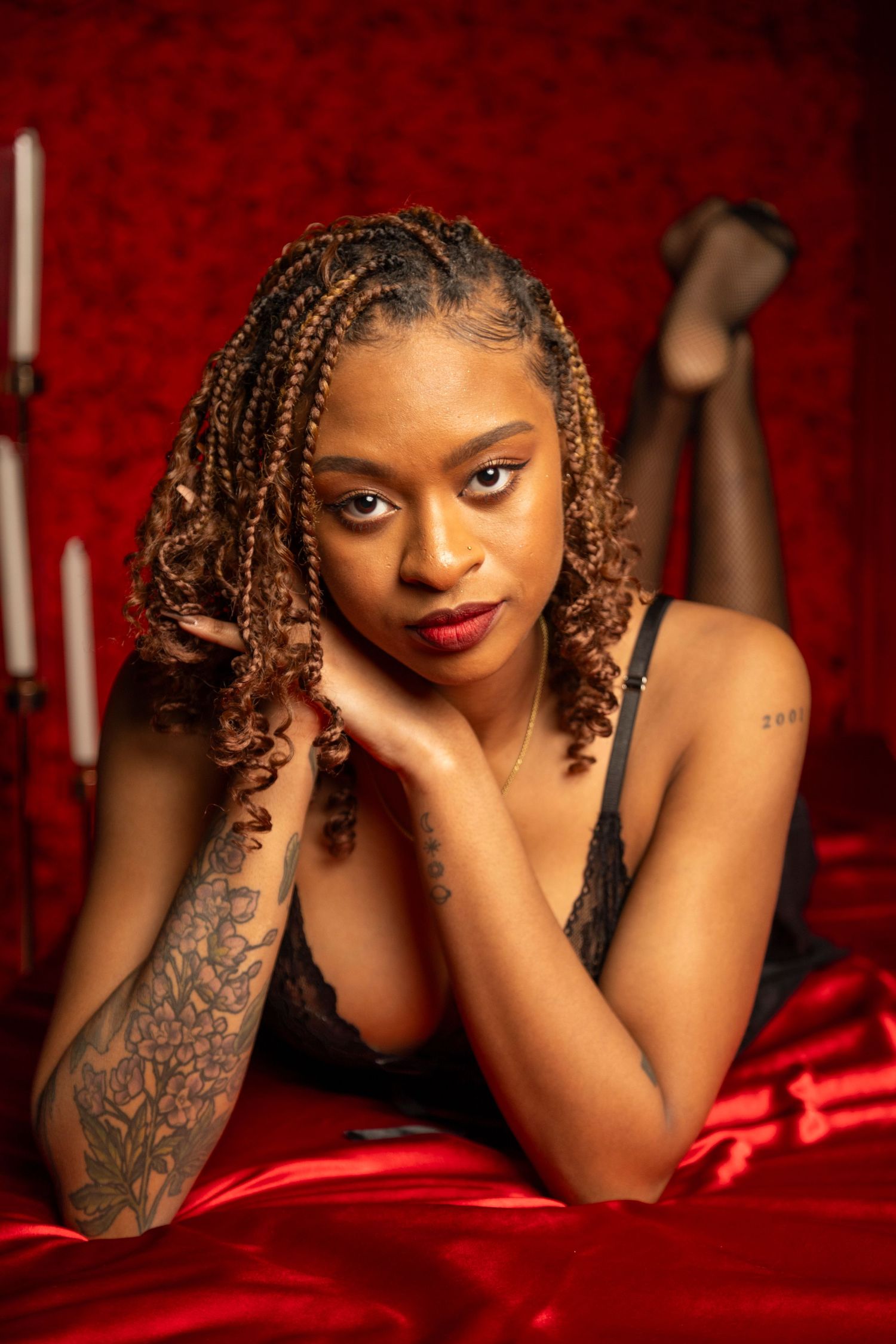 Young Black woman with braids and tattoos poses on red satin bed in black lingerie with candles in background.