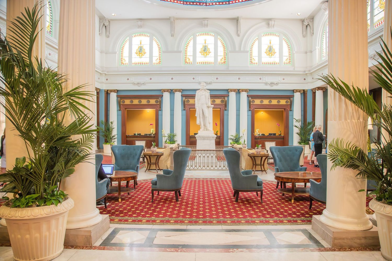 Elegant hotel lobby with blue armchairs, red patterned carpet, white columns, and tropical plants in a grand classical interior.