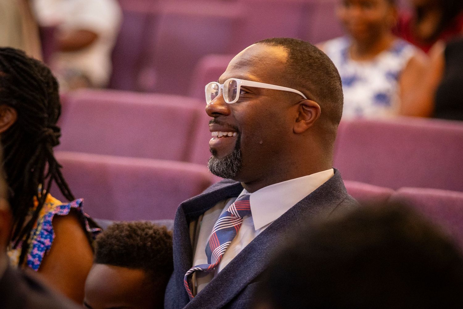 Smiling man in suit and white glasses seated in purple auditorium chairs at a formal event.