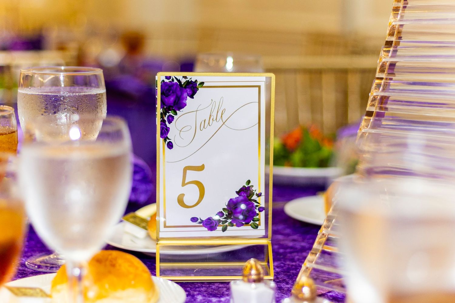 Purple floral table number 5 card in gold frame at elegant wedding reception with purple tablecloth.