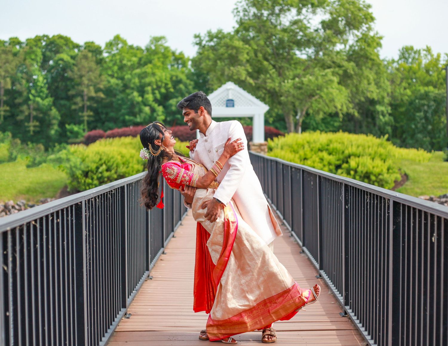 A couple in traditional Indian attire walks along a wooden bridge surrounded by lush greenery.
