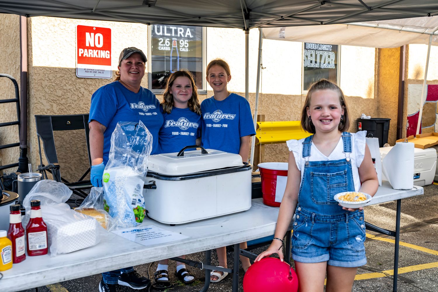 Volunteers in blue shirts staff refreshment tables under a white canopy tent at a community event.