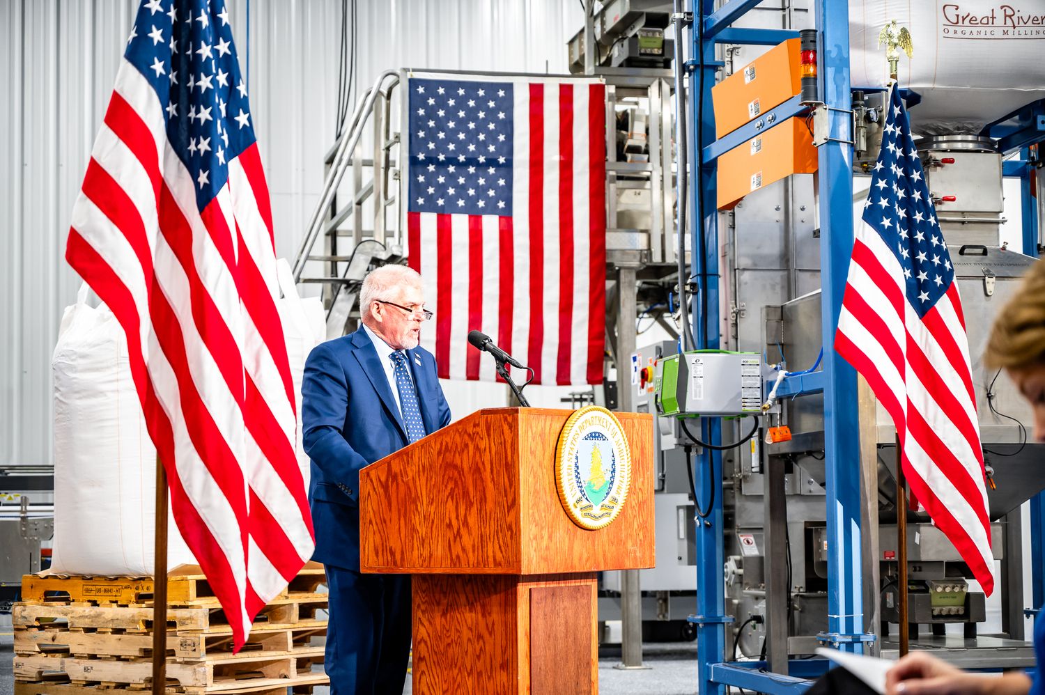 Official ceremony setup with American flags and podium in an industrial warehouse setting.
