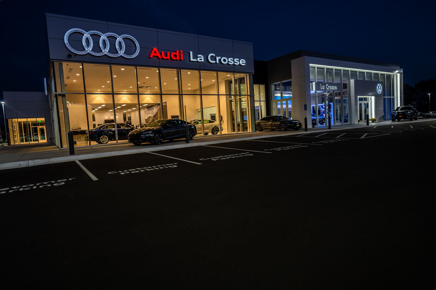 Illuminated Audi La Crosse dealership exterior at night with glass showroom windows displaying luxury vehicles.