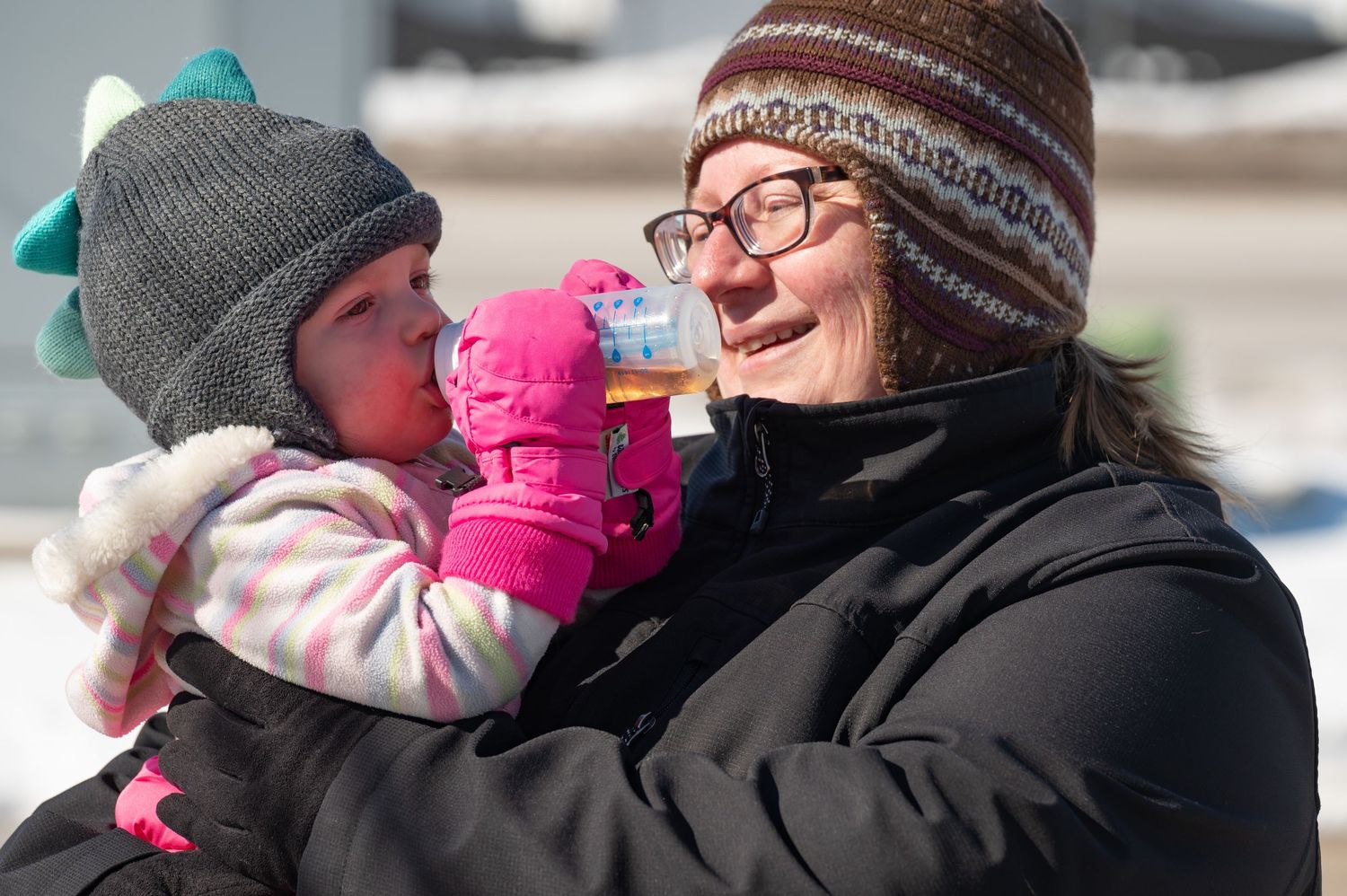 Woman in winter hat holds a baby drinking from a bottle outdoors in snowy weather.