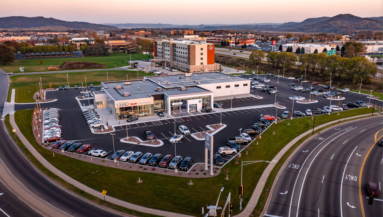 Bird's eye view of car dealership complex with curved access road and full parking lot at sunset.
