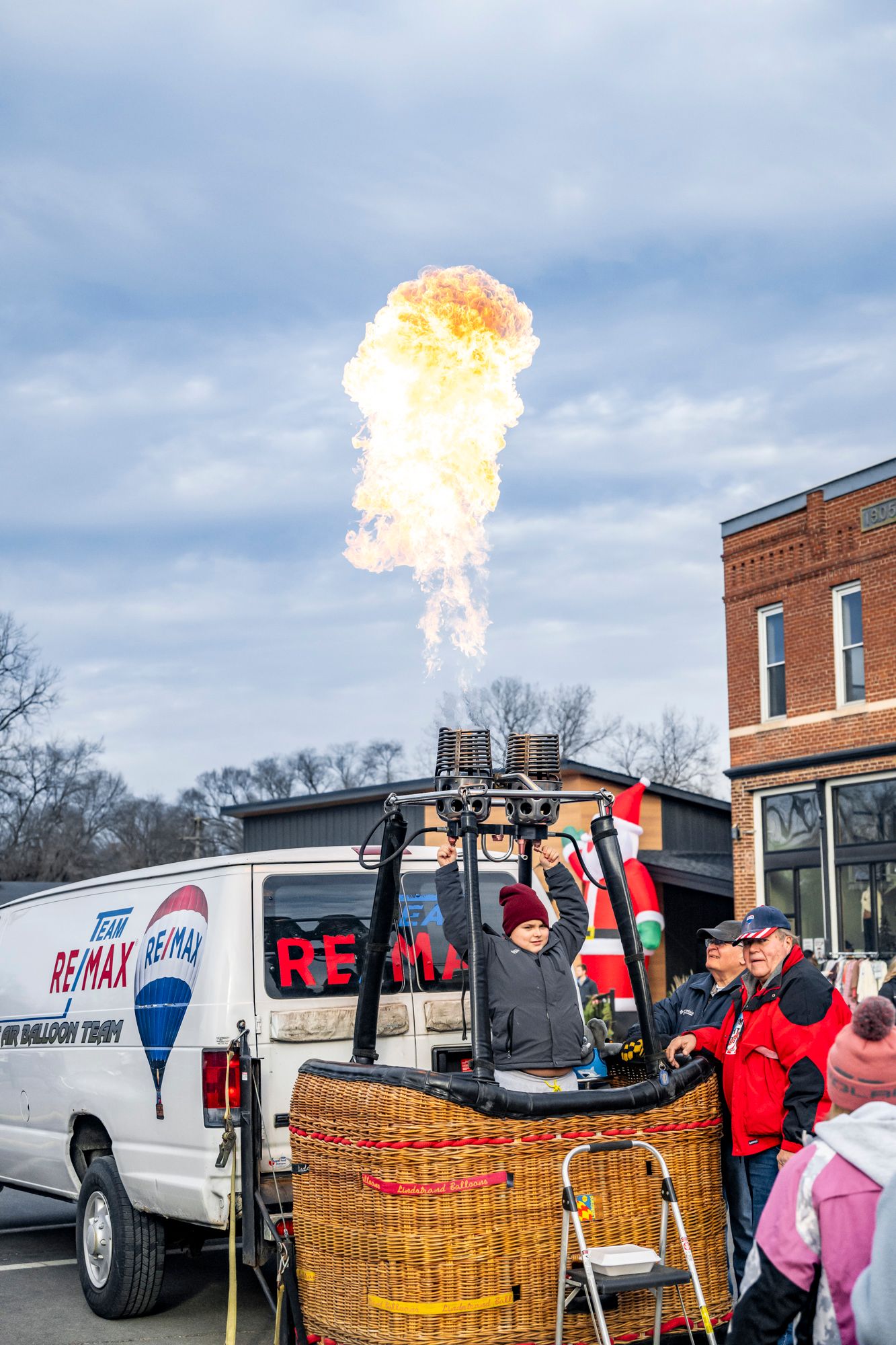 Hot air balloon demonstration with flames shooting upward near parked vans and crowd.