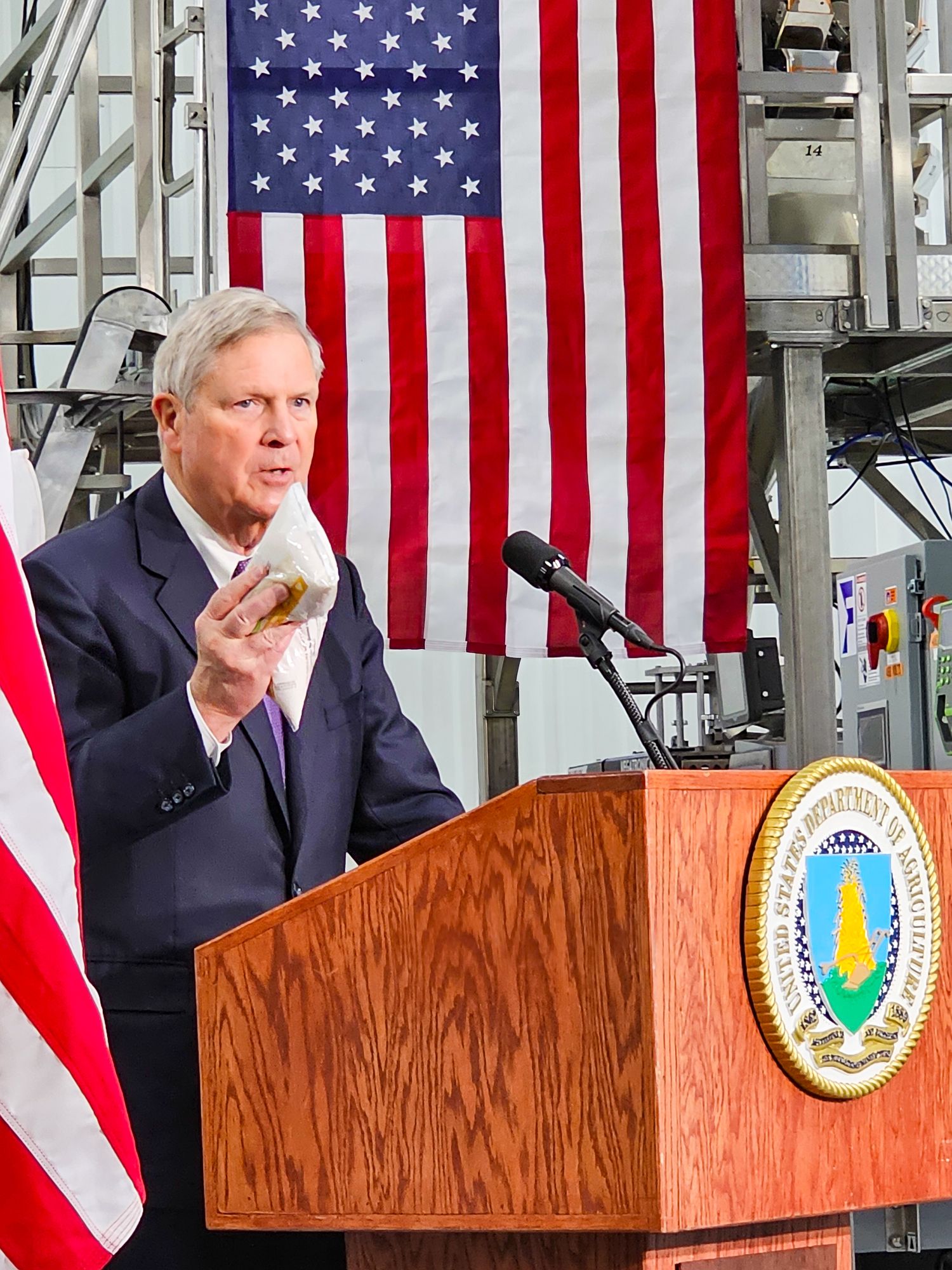Speaker at wooden podium with Department of Agriculture seal and American flags in background.