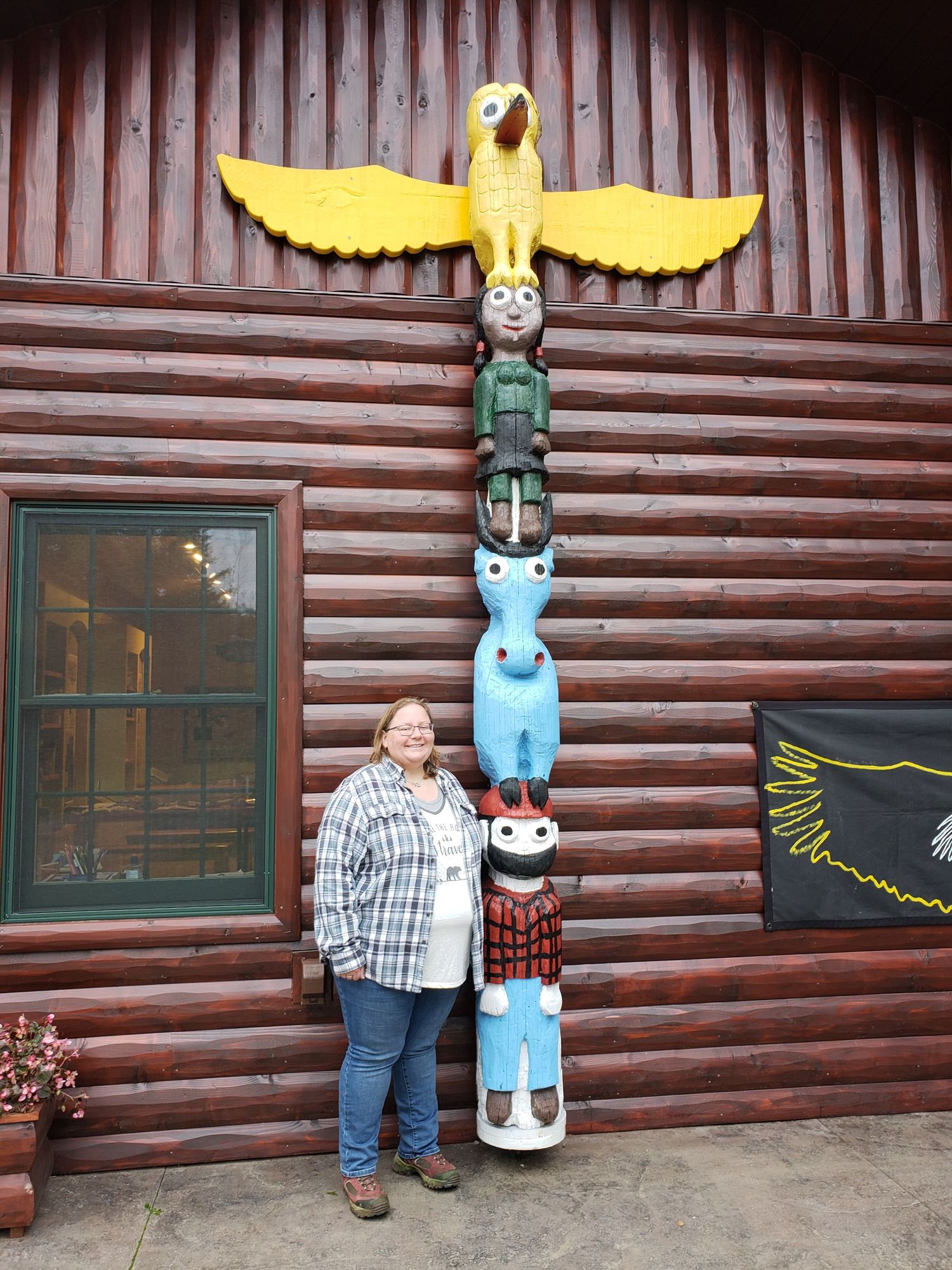 Woman posing beside a colorful totem pole with yellow bird on top, in front of a rustic log cabin building.