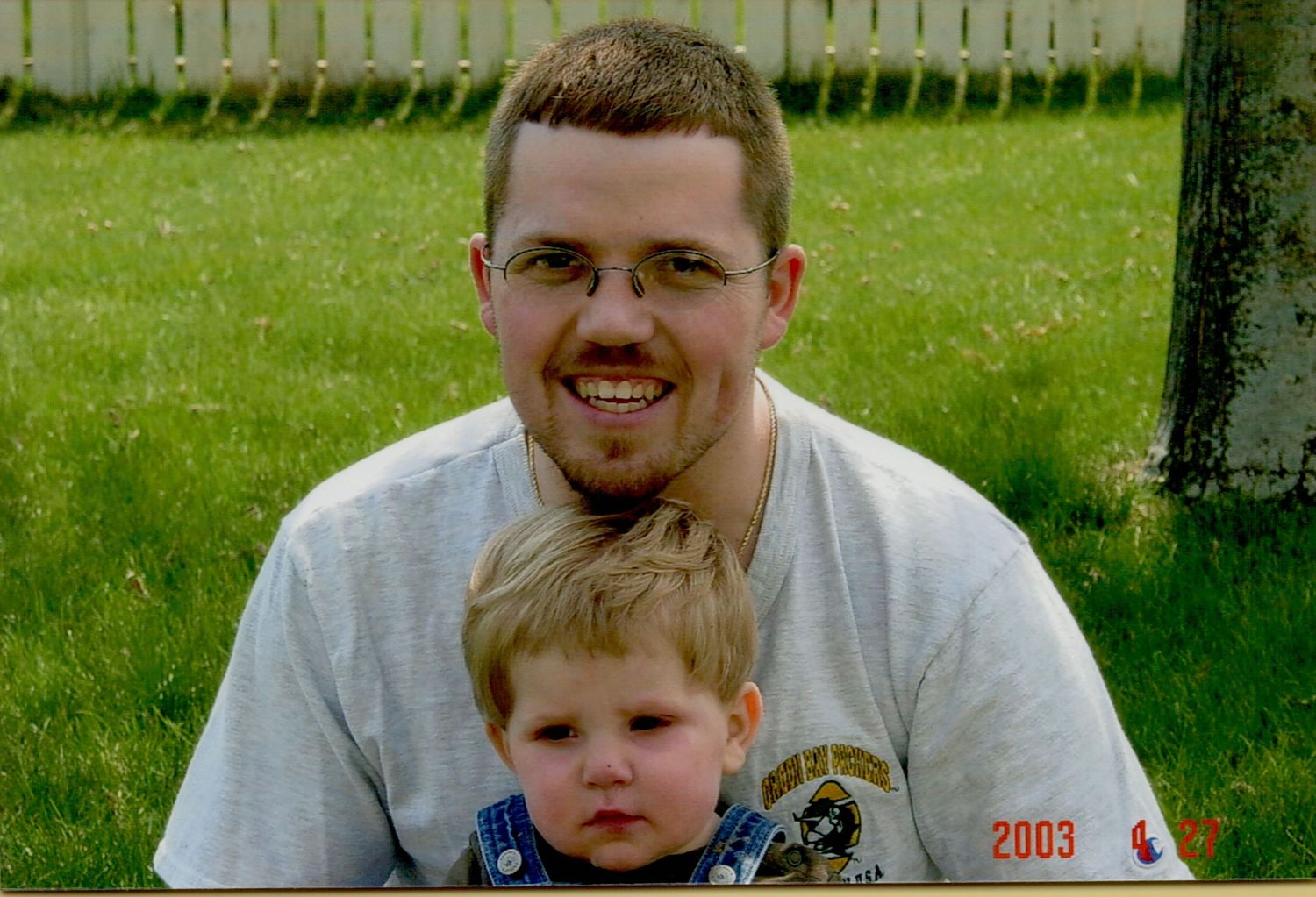 Man with glasses and goatee poses with young toddler boy outdoors on green lawn in 2003 photo.