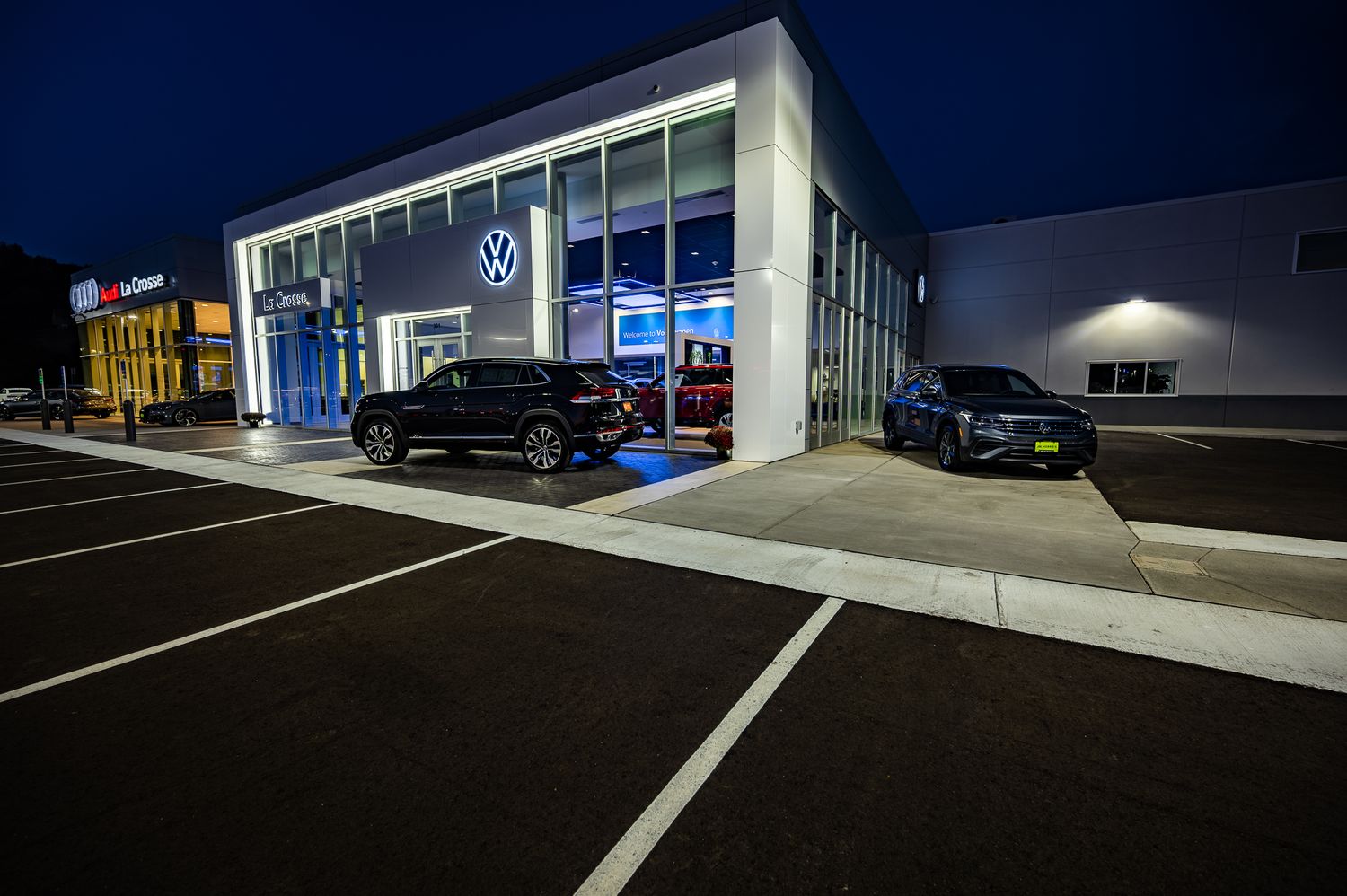Modern Volkswagen dealership exterior at night with illuminated showroom and parked vehicles.