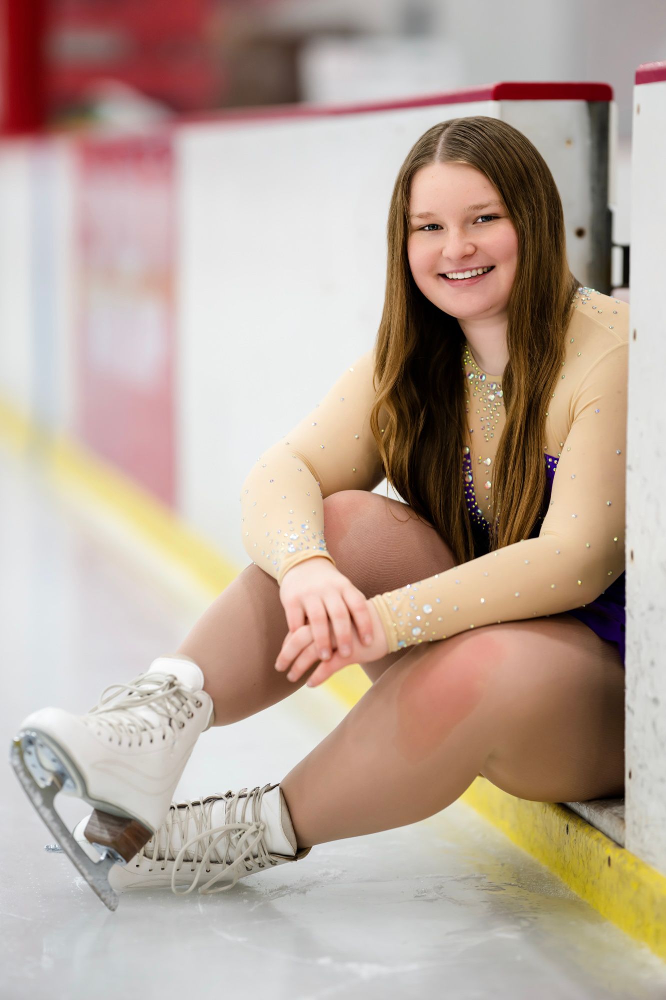 Figure skater sits on ice wearing white skates and purple performance attire.