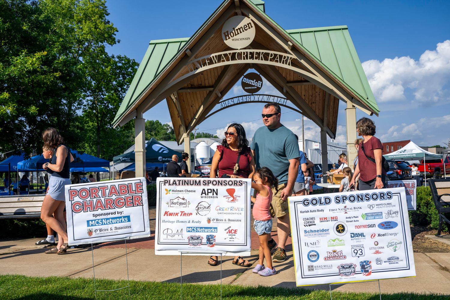 Row of promotional banners and signs displayed under a wooden pavilion at an outdoor event.