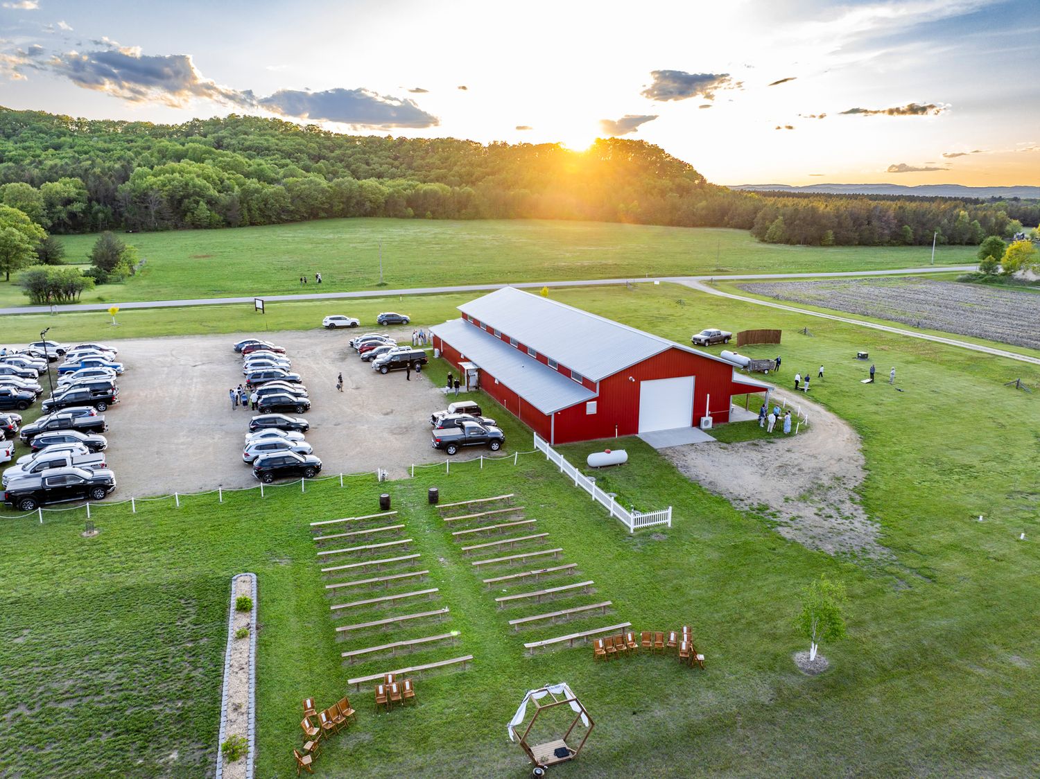Aerial view of a red barn and parking area during sunset with rows of chairs set up for an outdoor event.