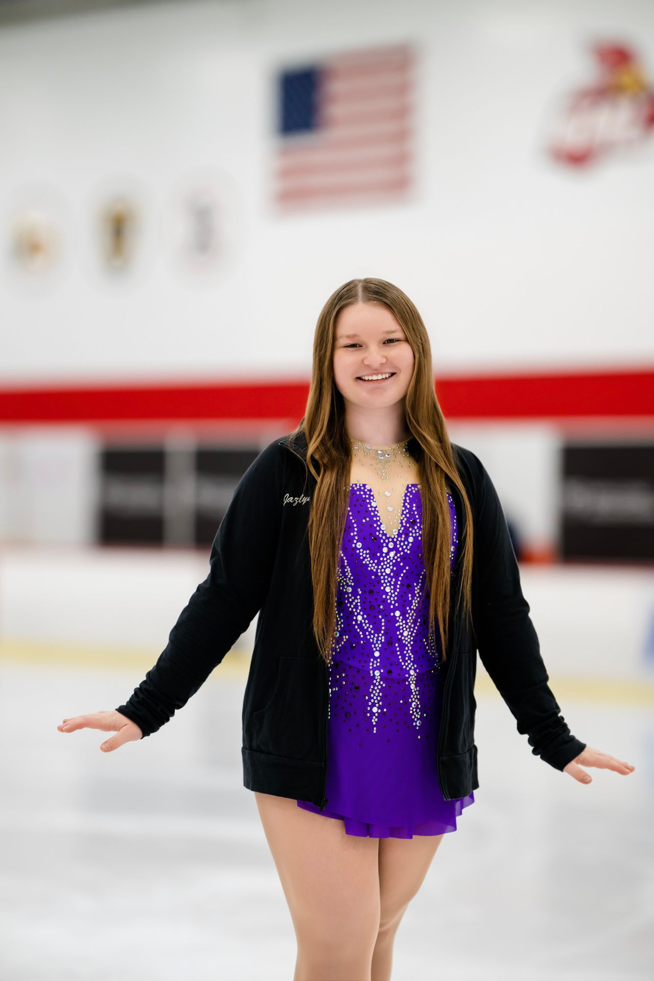 Skater in purple dress and black jacket poses on ice rink with American flag in background.