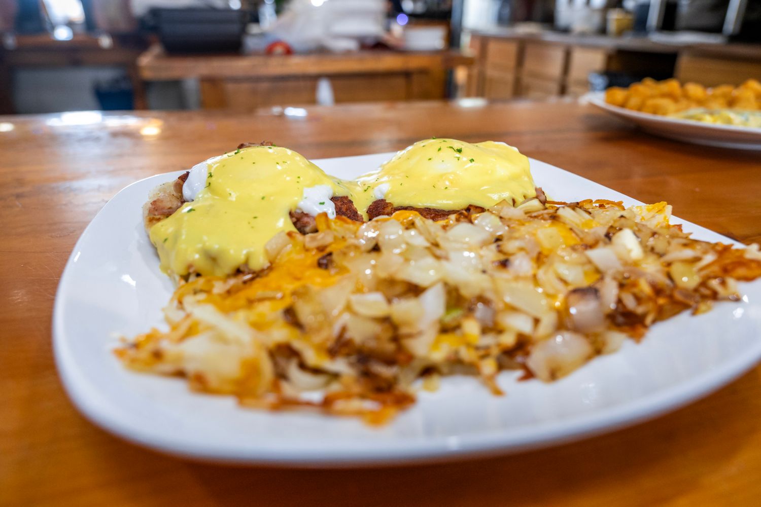 Eggs Benedict served with crispy hash browns on a white plate at a restaurant.