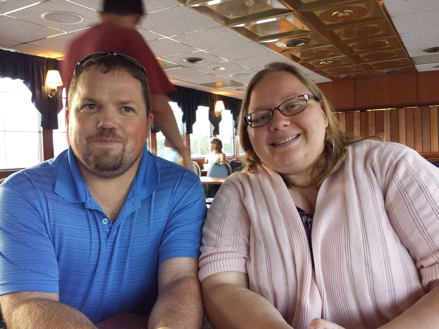 A man in a blue polo shirt and a woman in a pink cardigan with glasses smile together at a restaurant table.