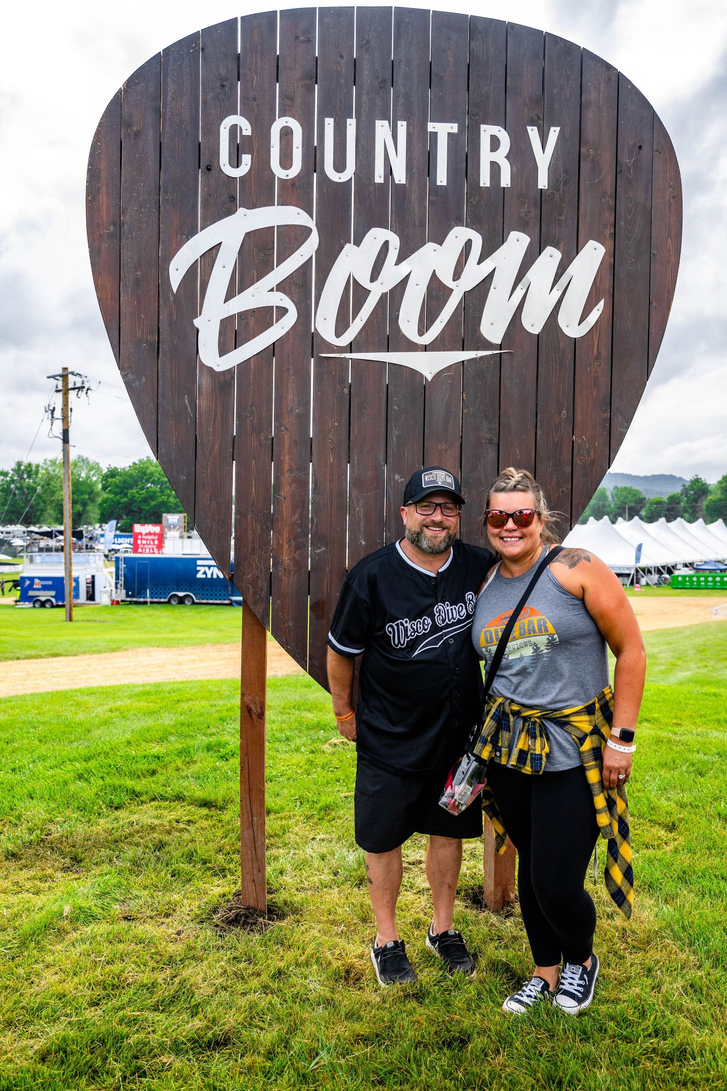 Two festival attendees pose in front of a large wooden guitar pick sign that says 'Country Boom' at an outdoor event.