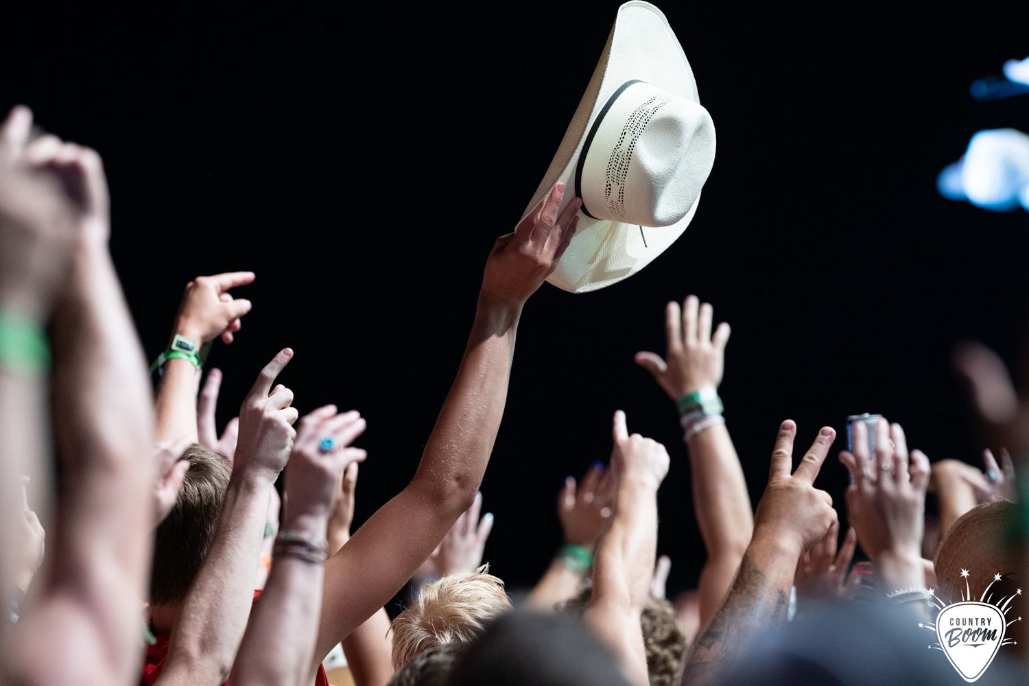 Crowd at outdoor concert with hands raised, one person holding a white cowboy hat above the crowd.