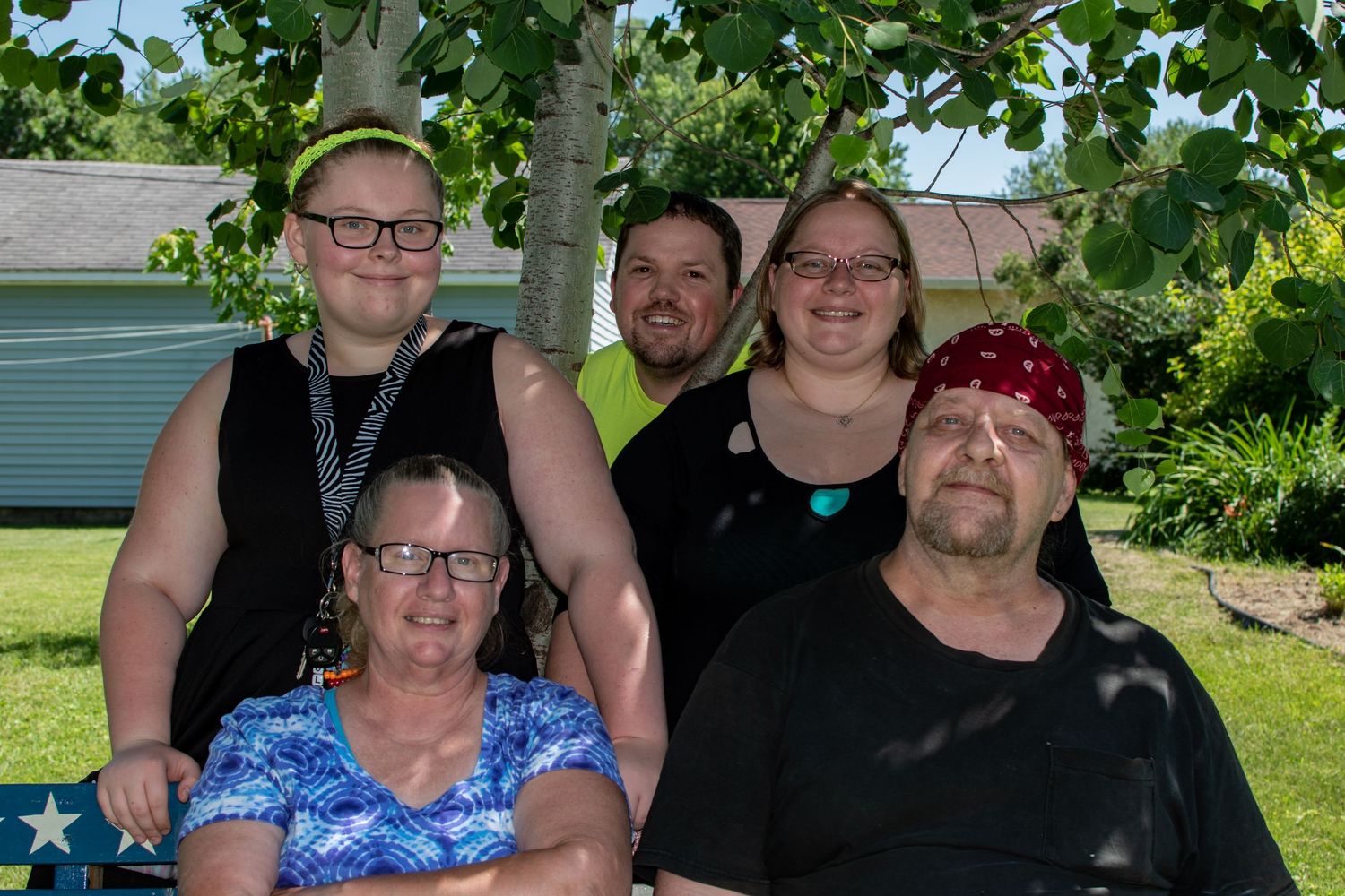 Family of five smiling outdoors near a birch tree on a sunny day in a green backyard setting.