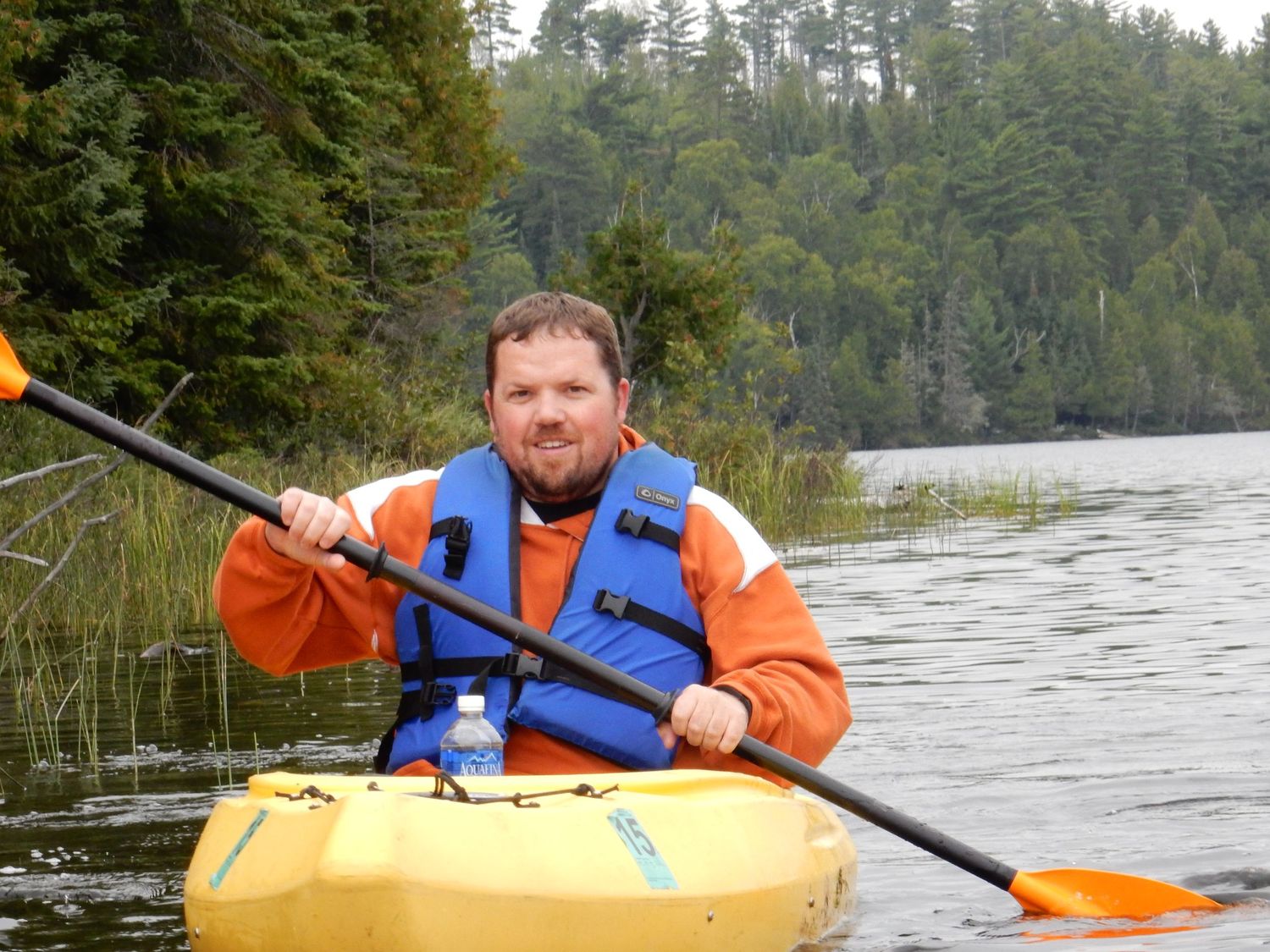 Man in orange jacket and blue life vest kayaking on calm lake surrounded by lush green forest.
