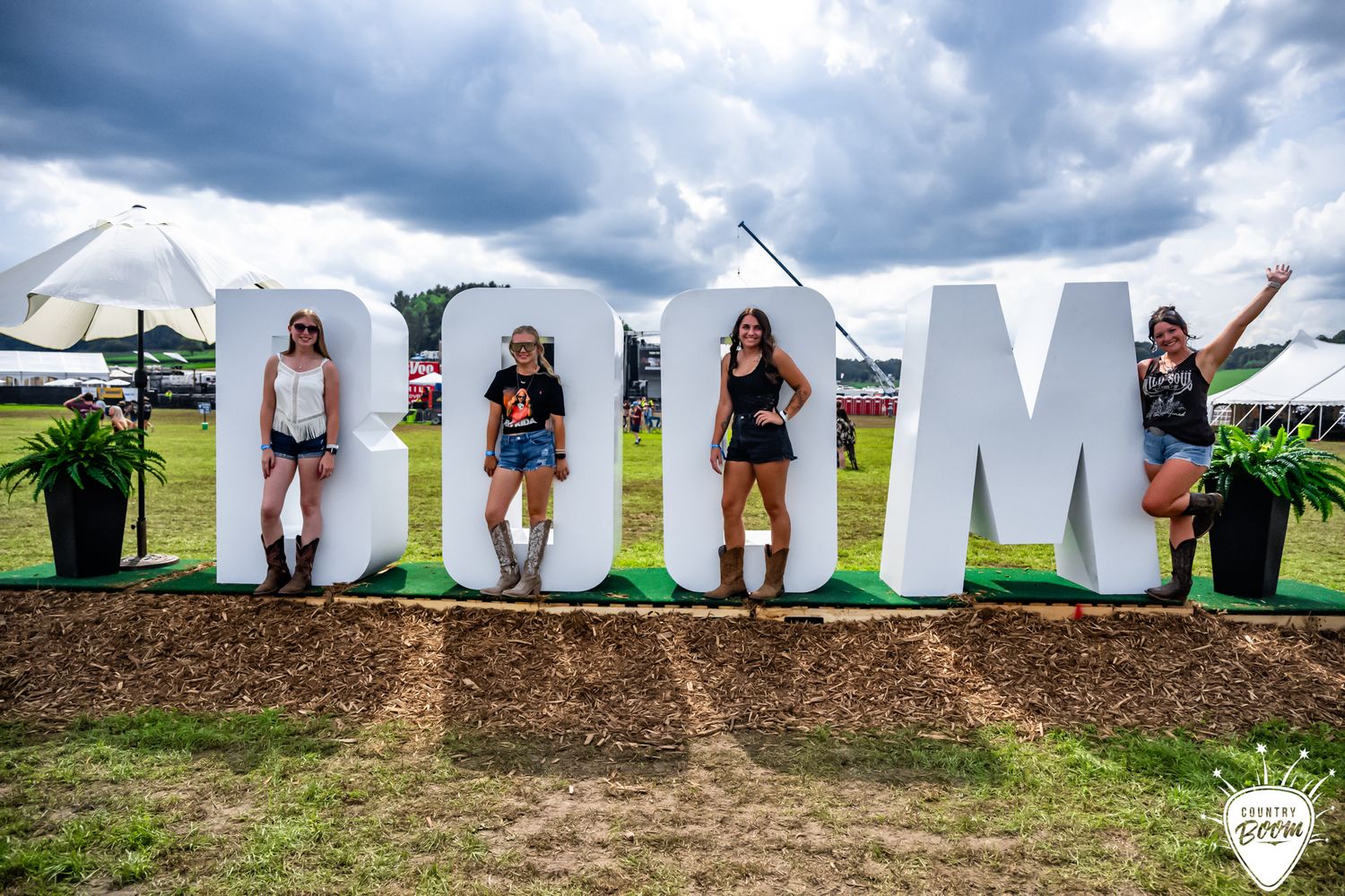 Three women pose in front of large white BOOM letters at an outdoor music festival on a cloudy day.