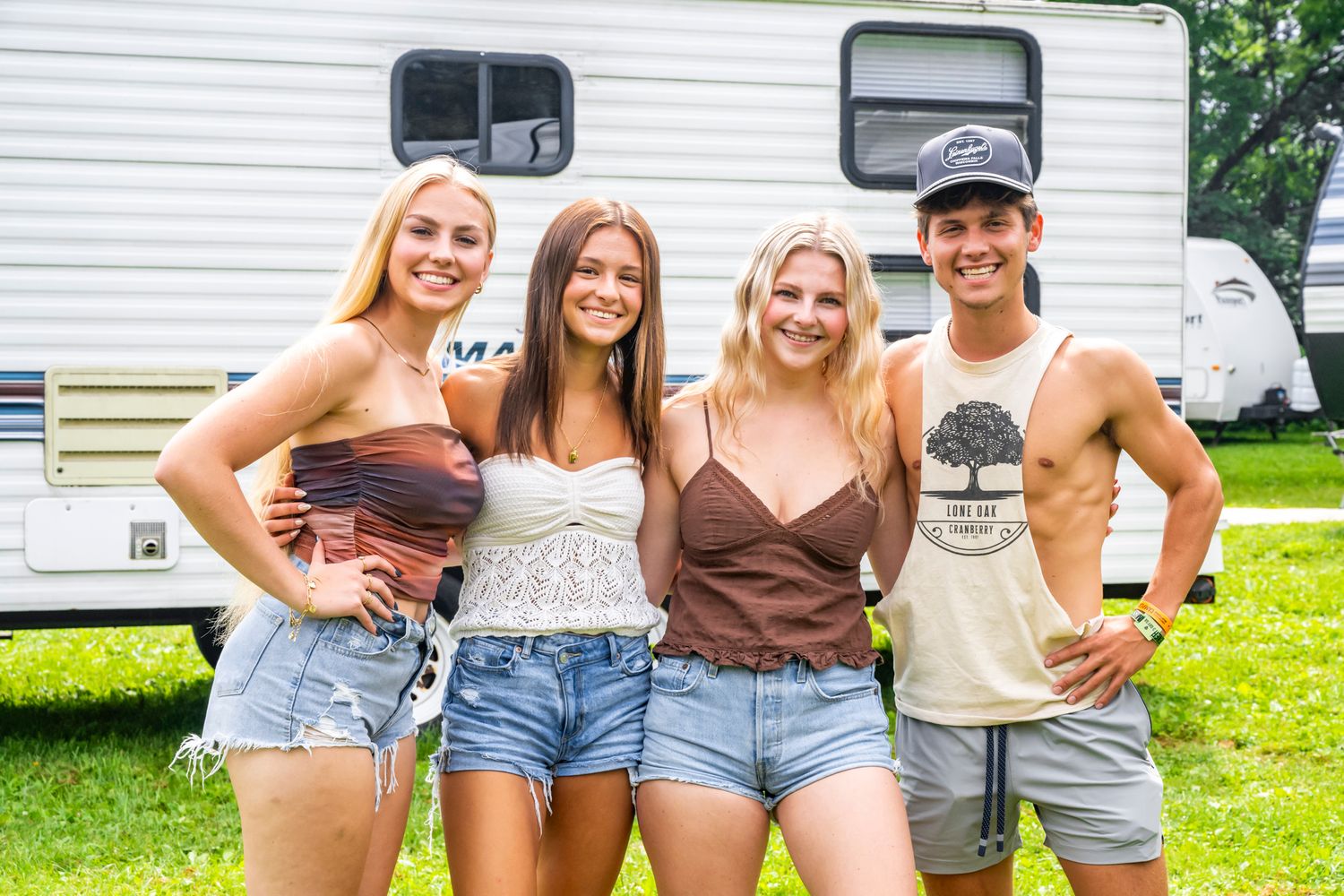 Group of friends pose together in casual summer outfits in front of a white RV at a music festival.
