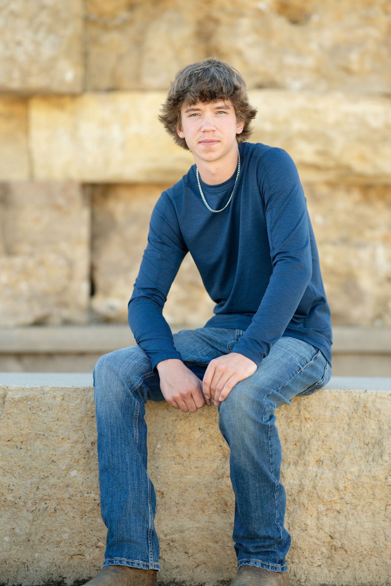 Casual portrait sitting on stone ledge wearing navy sweater and jeans.