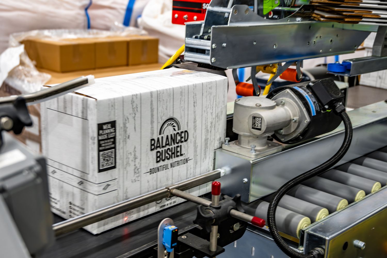 Industrial conveyor belt system moving Balanced Bushel branded boxes through a processing facility.