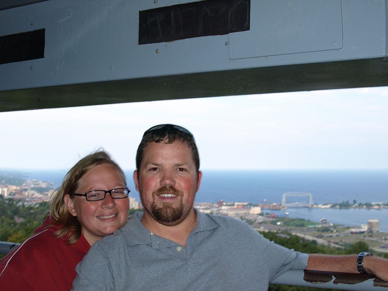 Couple smiling together at scenic overlook with harbor and city views in background on a clear day.
