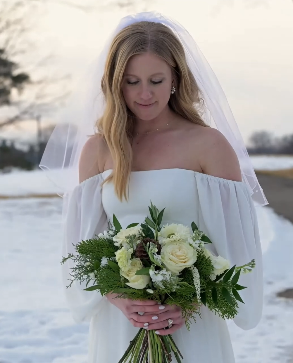 Bride in off-shoulder white dress holding winter white and green floral bouquet against snowy backdrop.