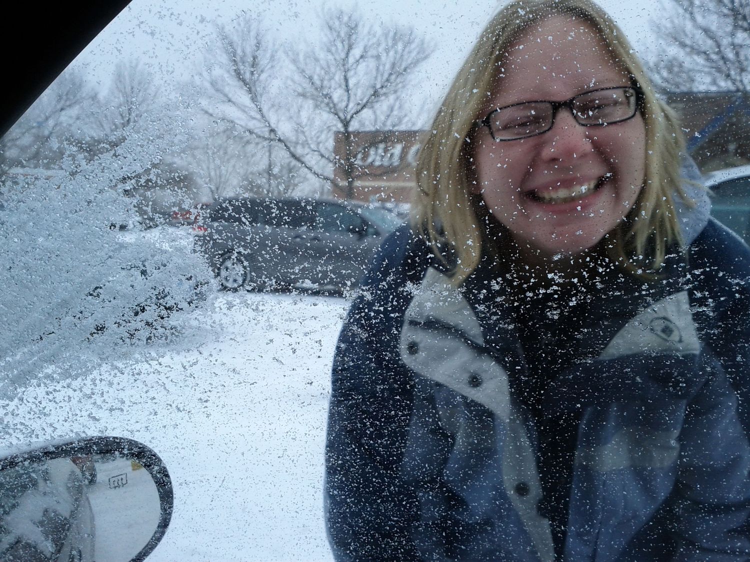 Smiling blonde woman with glasses stands outside in heavy snowfall near parked cars in winter.