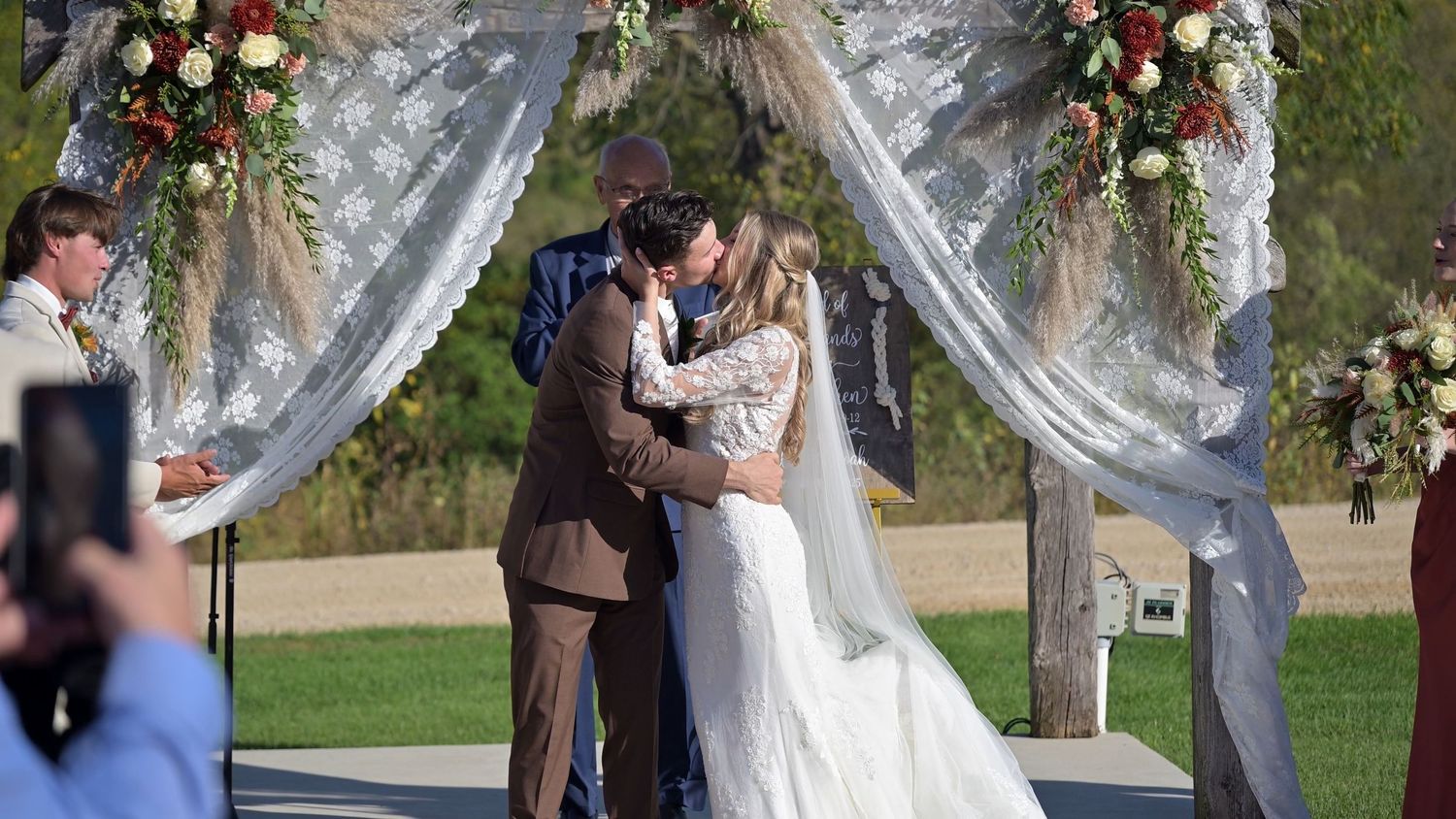 Wedding ceremony taking place under a white floral arch with guests seated outdoors.