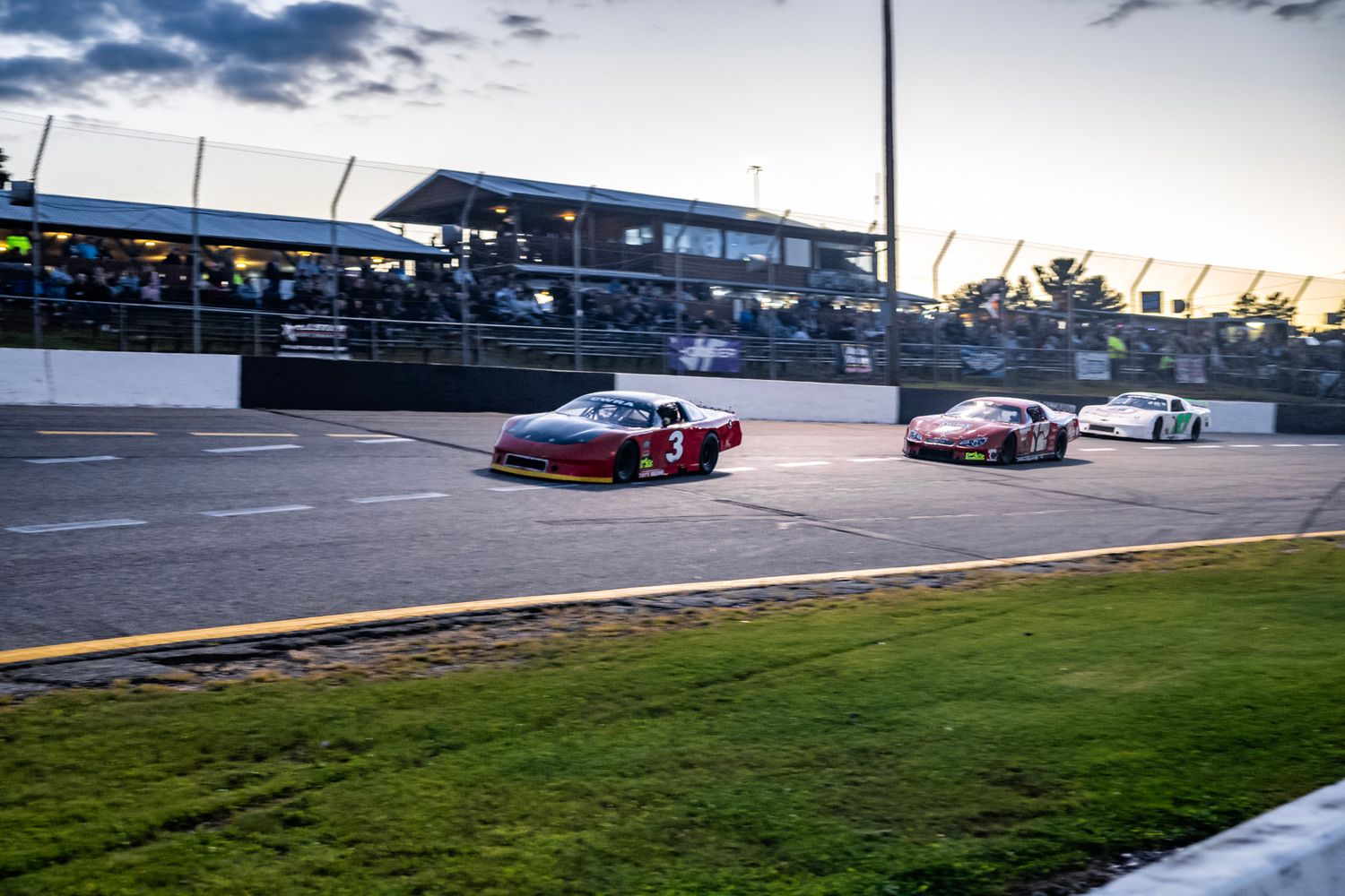 Race cars line up on track against dramatic cloudy sky at dusk.