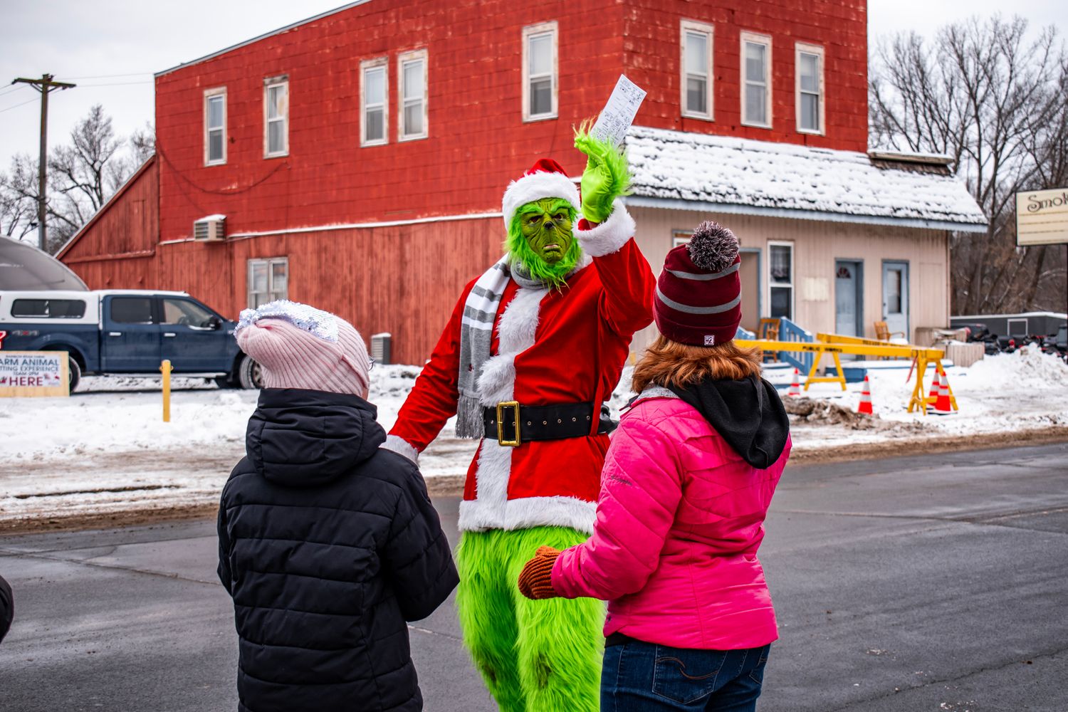 Character in green costume dressed as holiday Grinch interacting with visitors on snowy street.