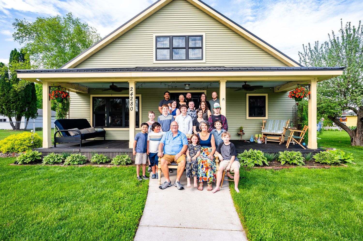 A large group gathered on the front porch of a craftsman-style home with beige siding and white trim on a sunny day.