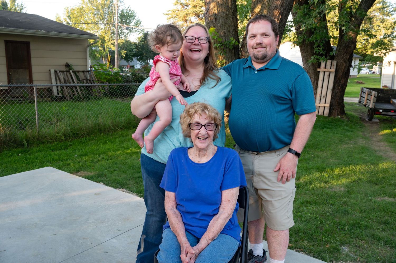 Family of four poses outdoors, including grandmother in wheelchair, couple, and young child, smiling together on a sunny day.