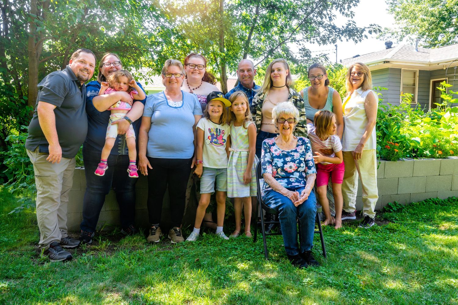 Extended family gathering outside traditional home with landscaped yard and greenery.