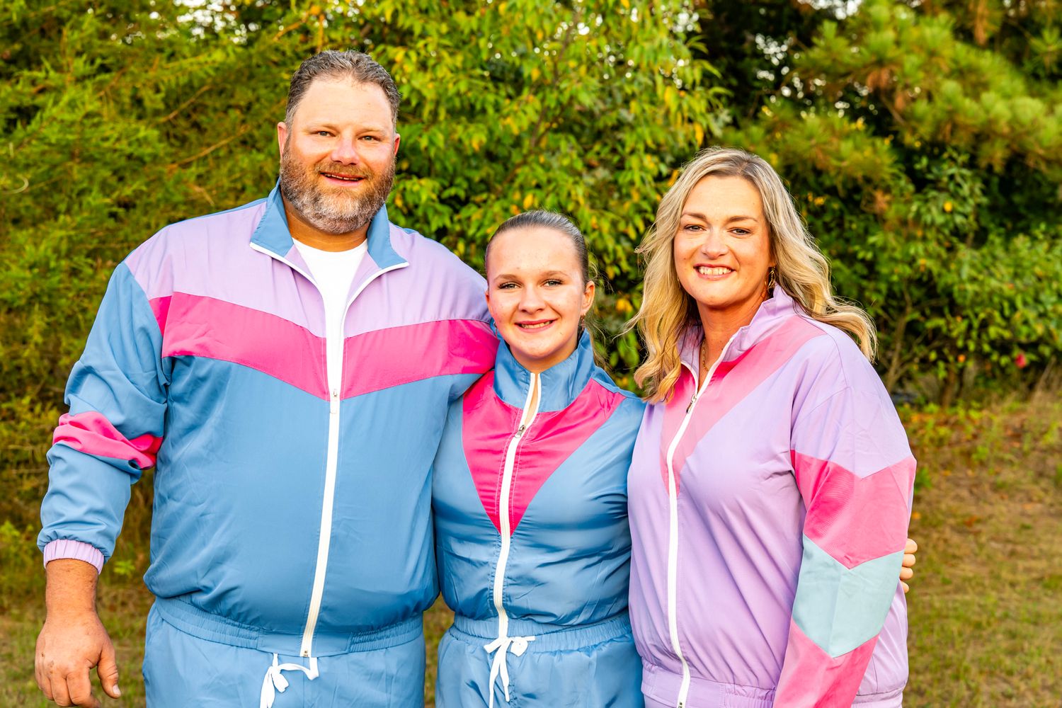 Three people wearing coordinated blue and pink windbreaker suits smiling outdoors.