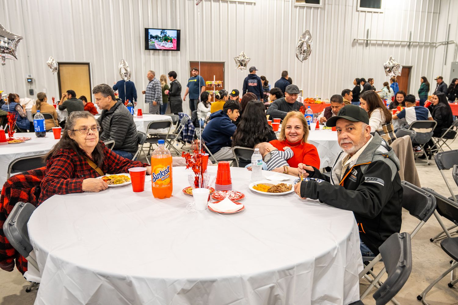 Community gathering in a large hall with people seated at round tables enjoying a meal together.