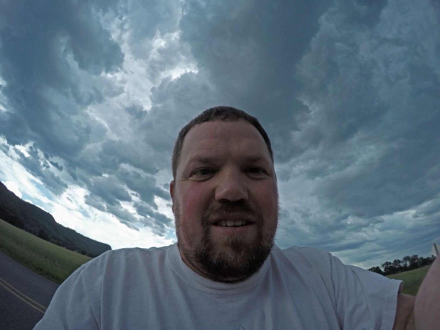 Man with beard in white t-shirt takes outdoor selfie under dramatic stormy cloudy sky near green fields.