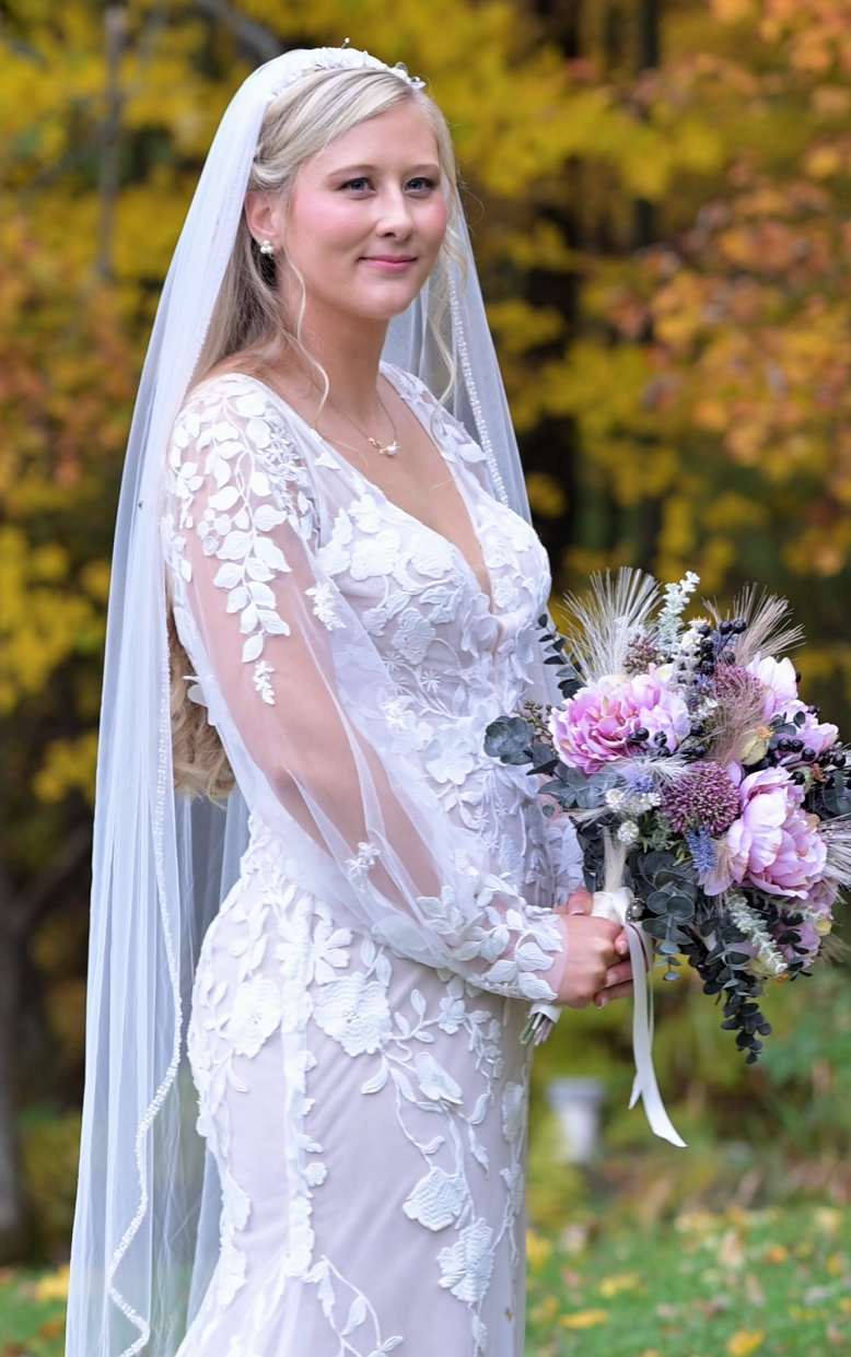 A bride in a detailed lace wedding dress poses with a pink and purple bouquet against autumn foliage.