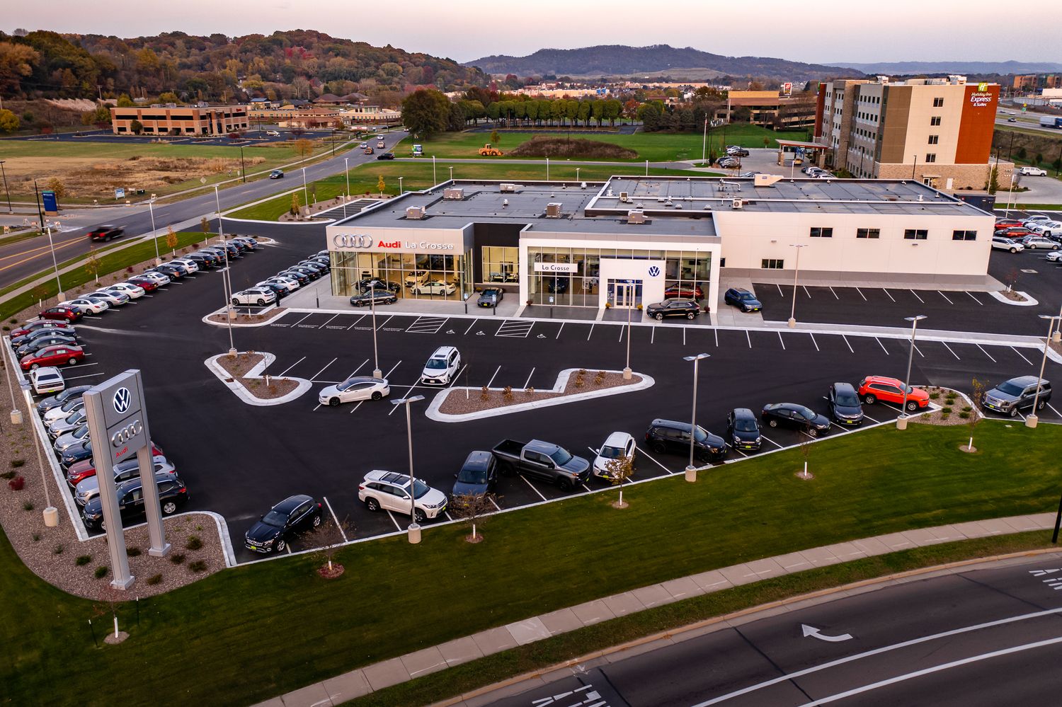 Aerial view of modern car dealership with large parking lot and surrounding landscape at sunset.