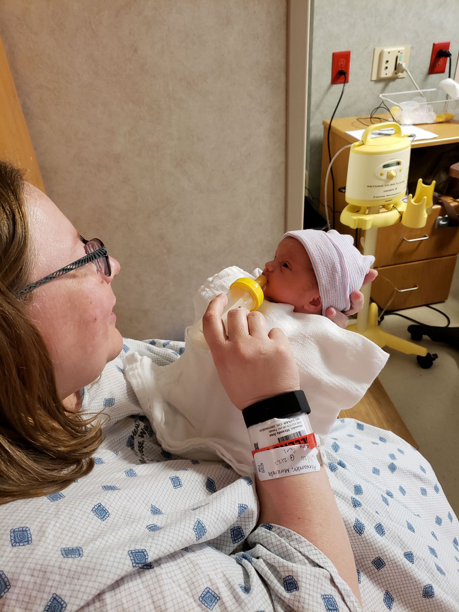 New mother in hospital gown bottle-feeding her newborn baby wearing a pink hat in a hospital room.