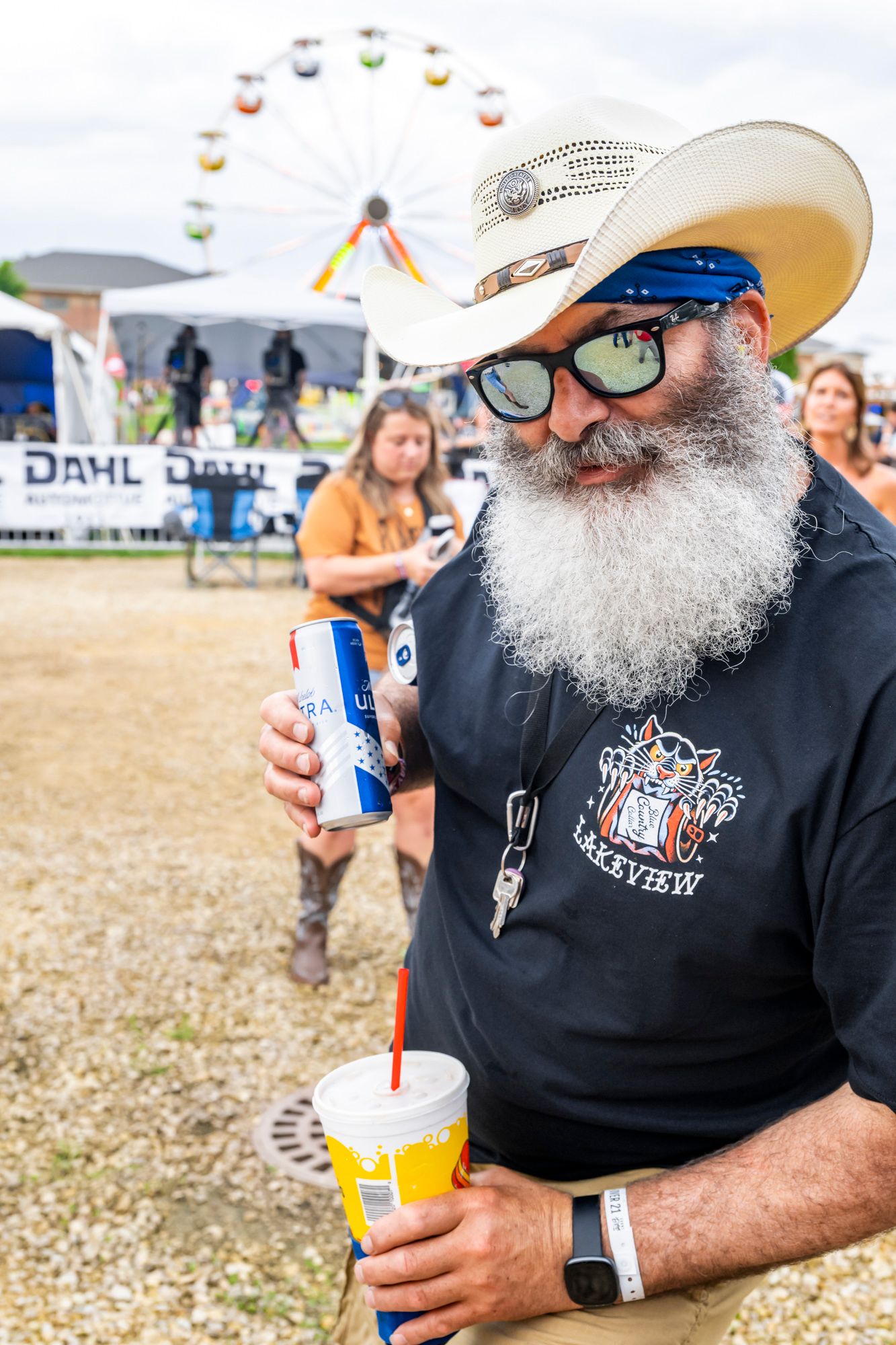 Man with white beard wearing cowboy hat and sunglasses holds beverages at outdoor festival with ferris wheel in background.