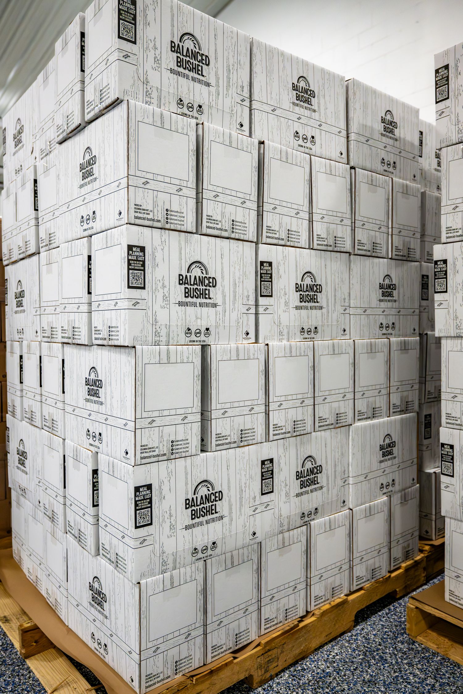 Stacked white cardboard boxes with Balanced Bushel branding in a warehouse storage area.