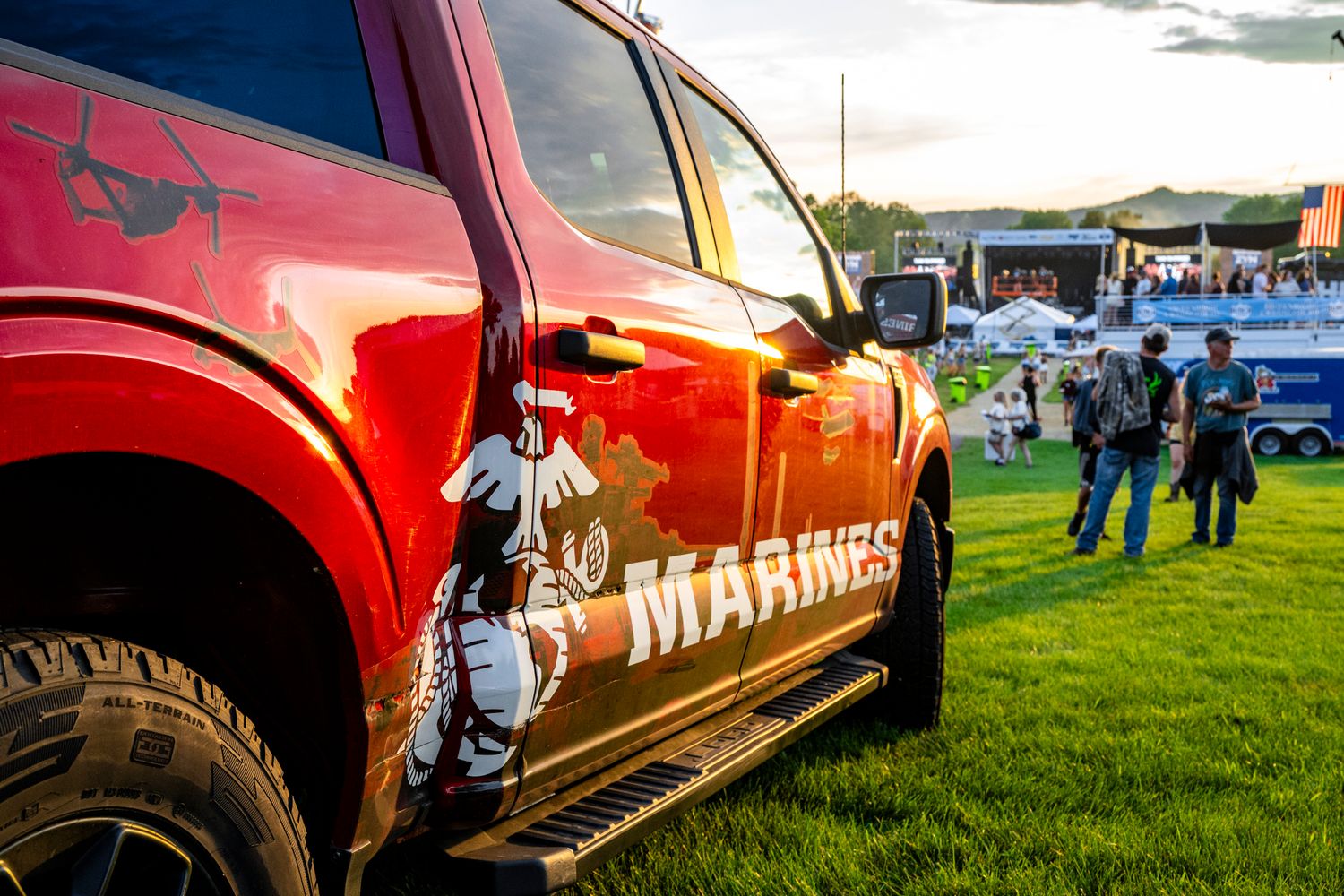 Red pickup truck with Marines branding parked on grass at outdoor festival event.